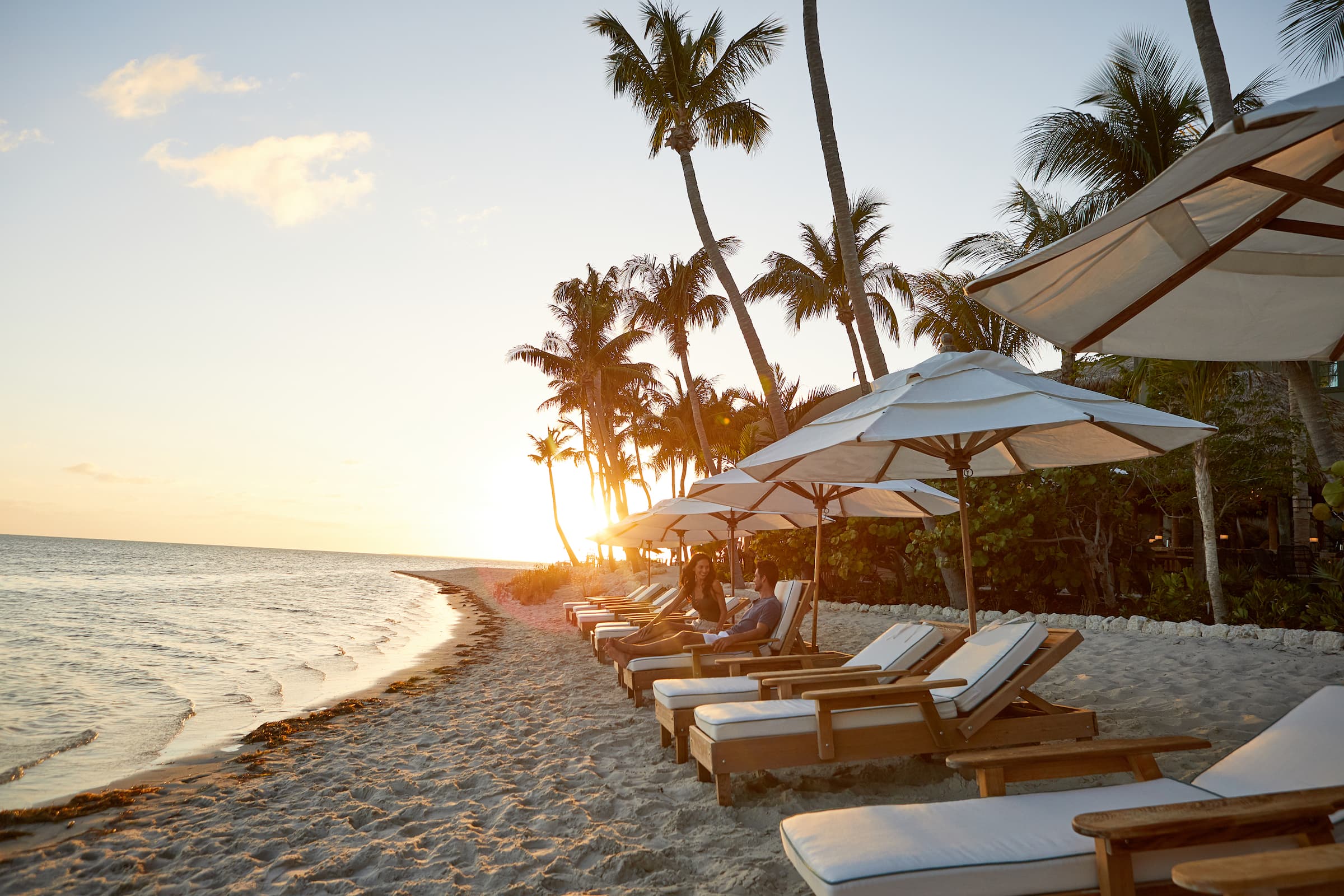 Wooden beach lounge chairs with white cushions on a sandy beach with the sun setting in the distance