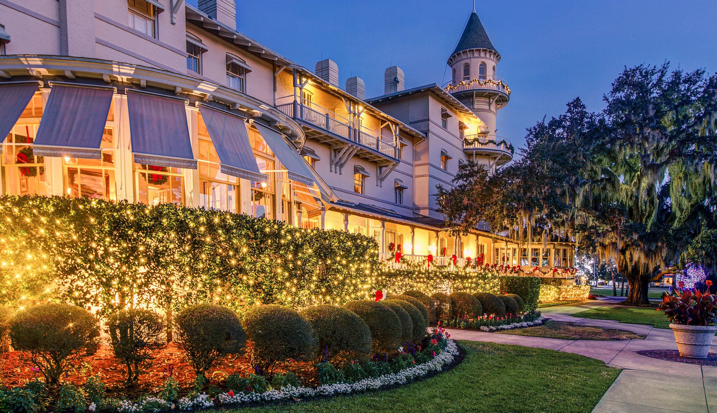 Holiday lights and festive garland illuminate the Victorian façade and turret of Jekyll Island Club Resort in Georgia at dusk.